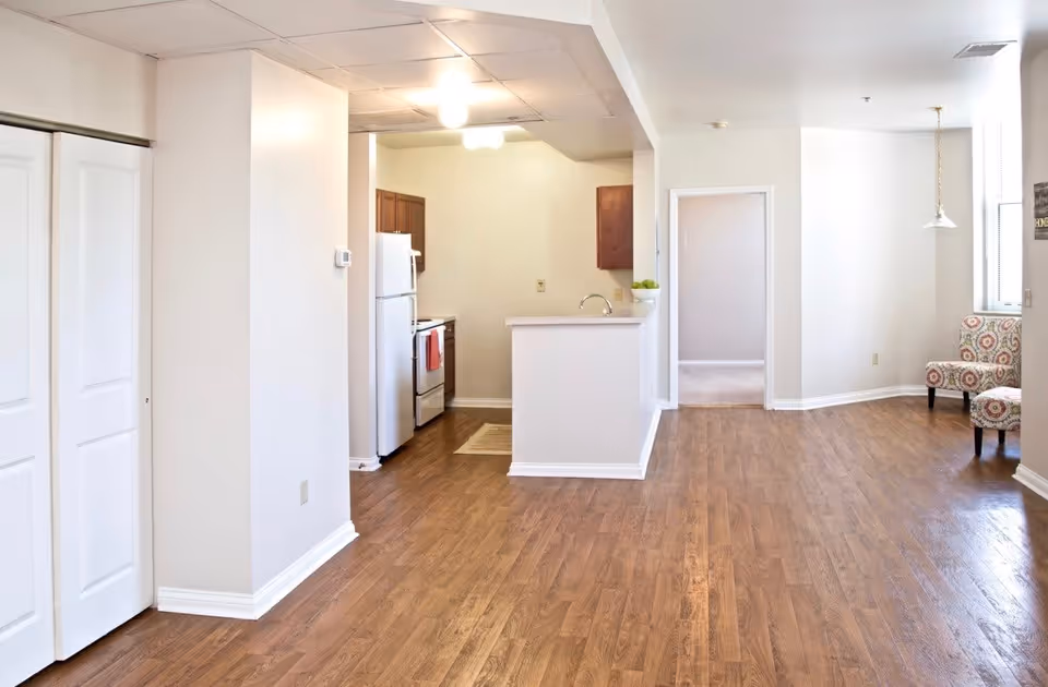 Interior view of a senior apartment showing a kitchen area with white refrigerator, stove, and wooden cabinets. The kitchen has a small counter with a sink and a bowl of green apples. The floor is wooden, and there is an open space leading to another room. Two patterned chairs are placed near a window with a hanging light fixture above.