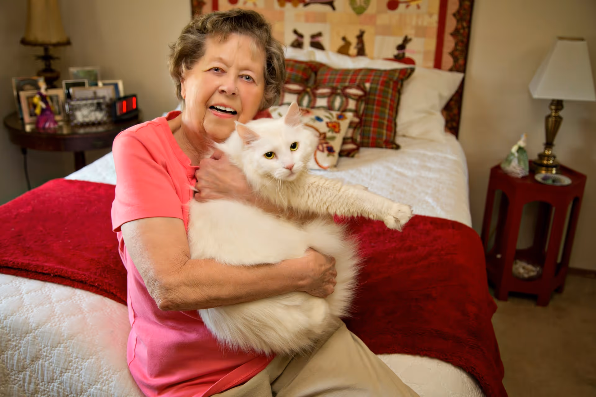 An elderly woman wearing a pink shirt sitting on a bed in a bedroom, holding a large white cat. The bed has a white quilt with a red blanket at the foot, and there are patterned pillows against the headboard. A nightstand with a lamp and decorative items is visible beside the bed.