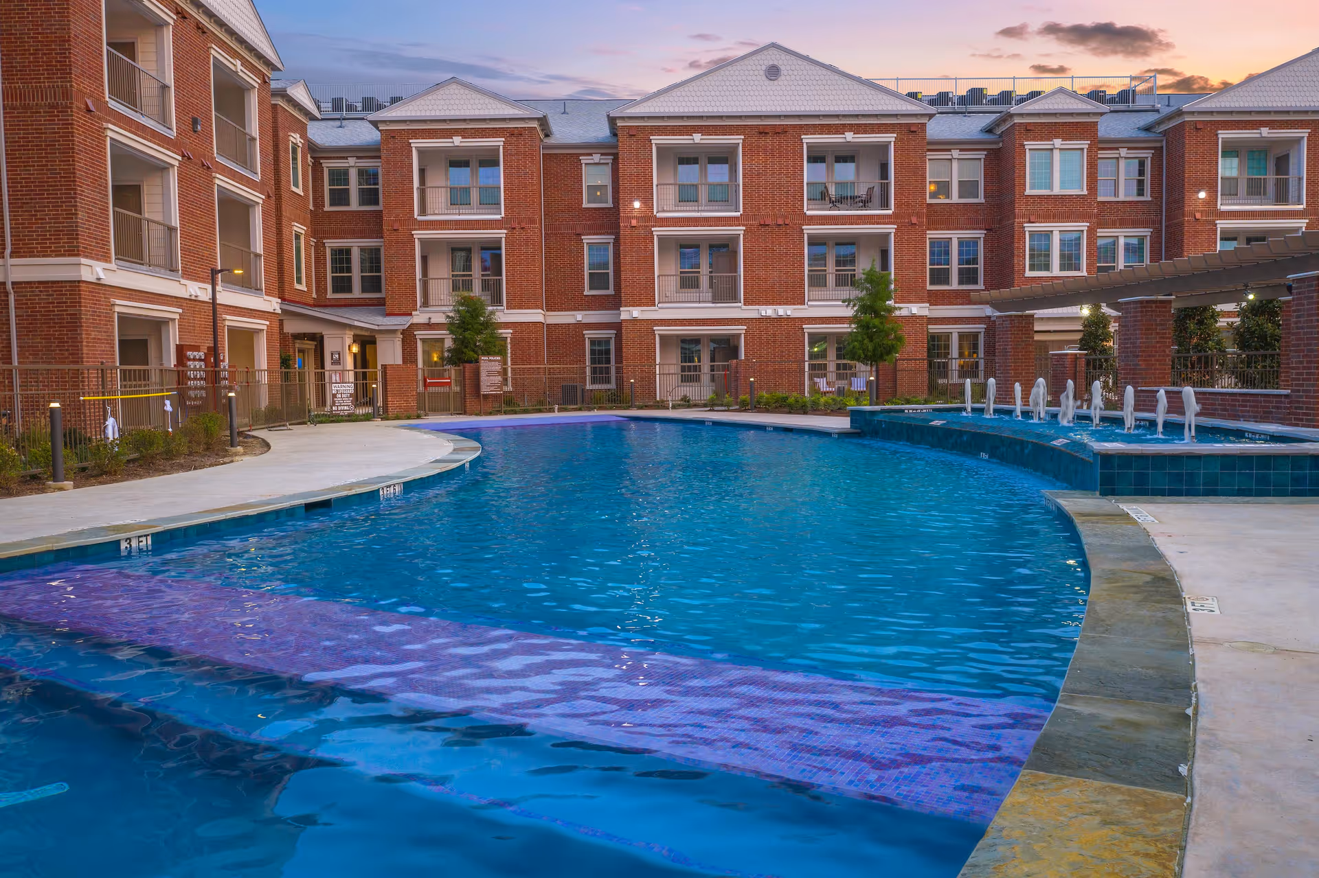 Outdoor swimming pool in a courtyard with a three-story red brick senior living building in the background at sunset.