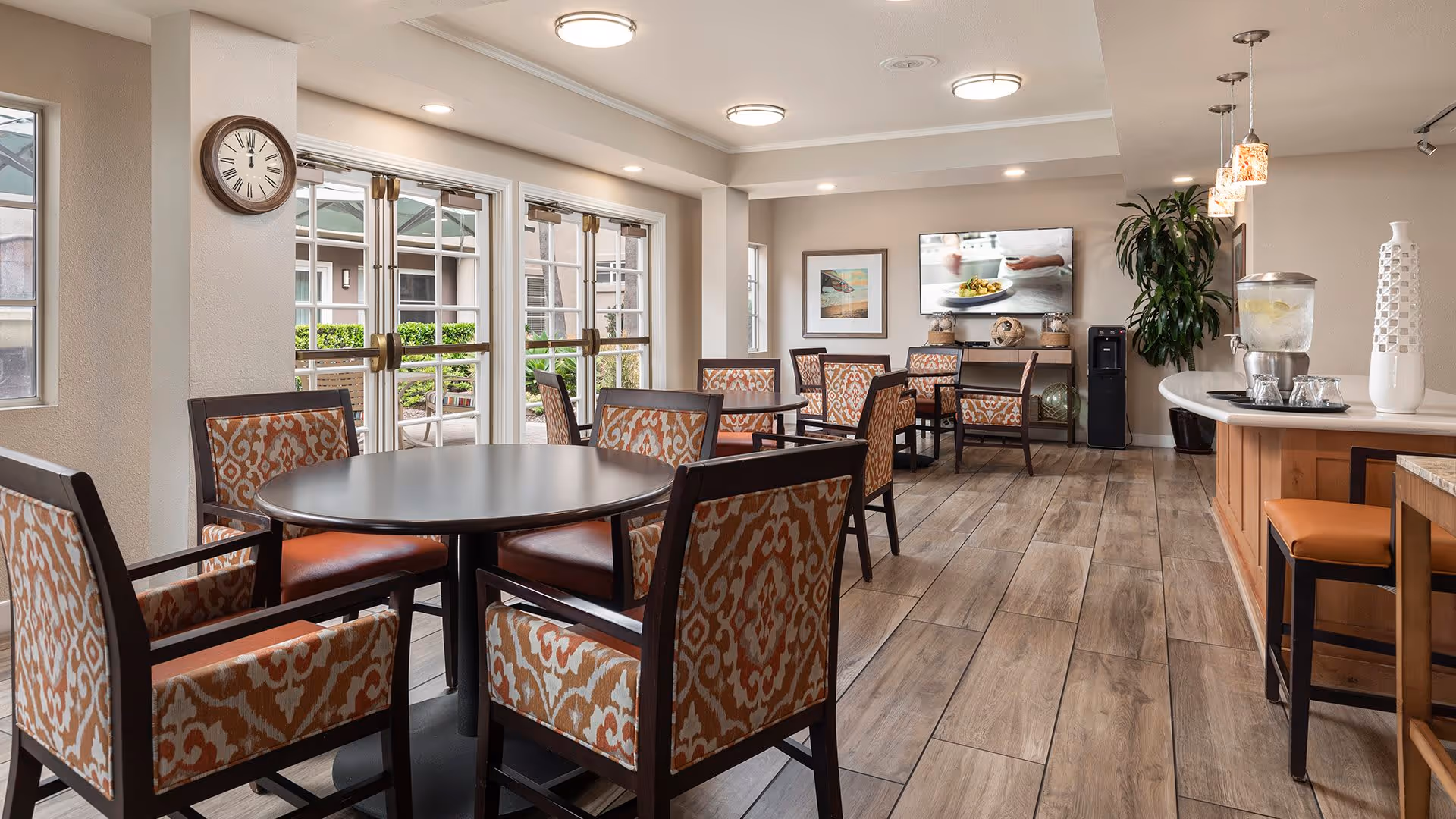A bright and spacious dining area with multiple round and square tables surrounded by patterned cushioned chairs. Large windows and glass doors allow natural light to fill the room. A clock is mounted on the wall, and a flat-screen TV is visible in the background above a console table. There is a counter with stools on the right side, decorated with a large vase and a water dispenser.