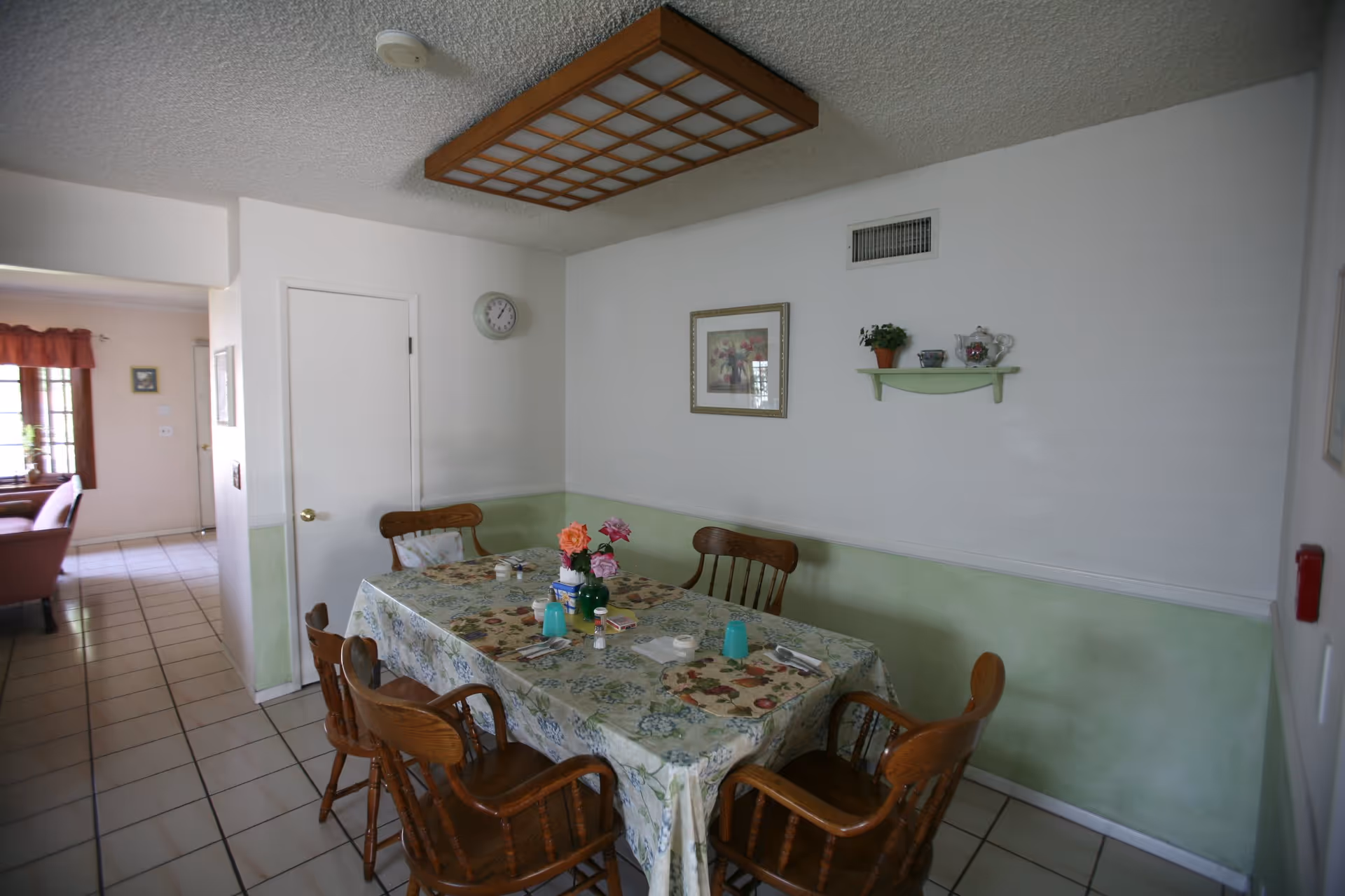 A dining area with a rectangular table covered with a floral tablecloth and set with placemats, utensils, salt and pepper shakers, and a vase of flowers. There are six wooden chairs around the table. The walls are painted white with a green lower half, and a framed picture and small shelf with decorative items hang on the wall. A clock is mounted on the wall near a closed door, and a large wooden light fixture is on the ceiling. The floor is tiled, and an adjacent room with a window and curtains is visible in the background.