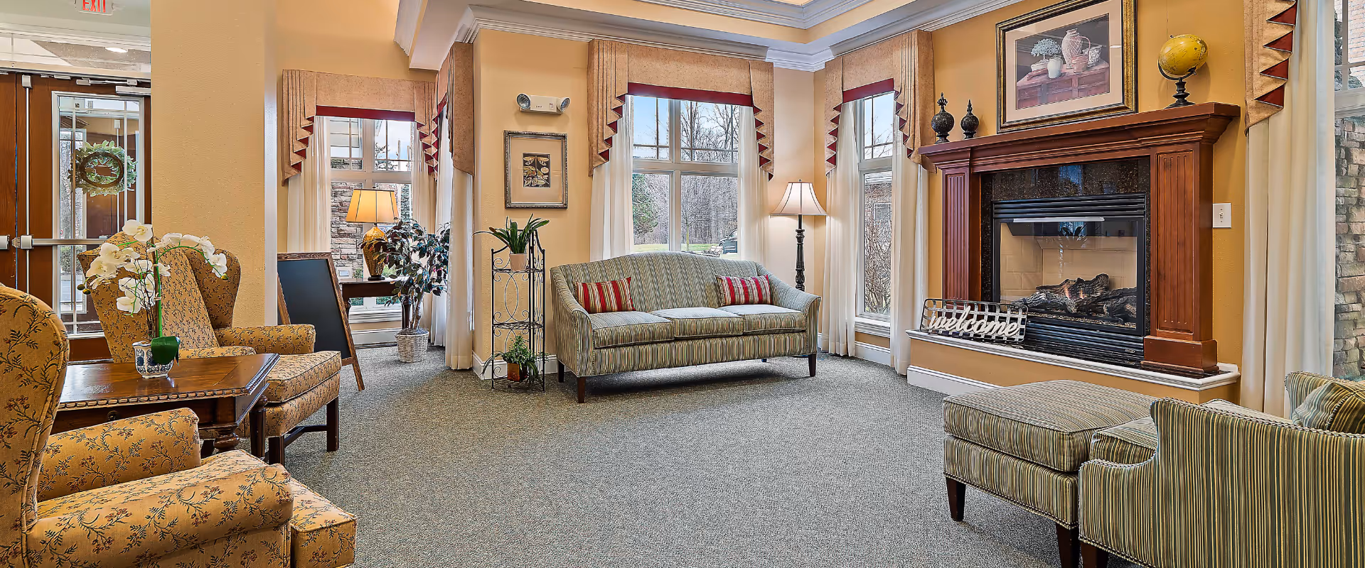 A cozy living room area with a fireplace, a striped sofa with red and white pillows, two patterned armchairs, a wooden coffee table with a flower vase, and large windows with curtains letting in natural light. There is a 'Welcome' sign on the fireplace mantel and framed artwork above it. The room has beige walls and carpeted flooring.