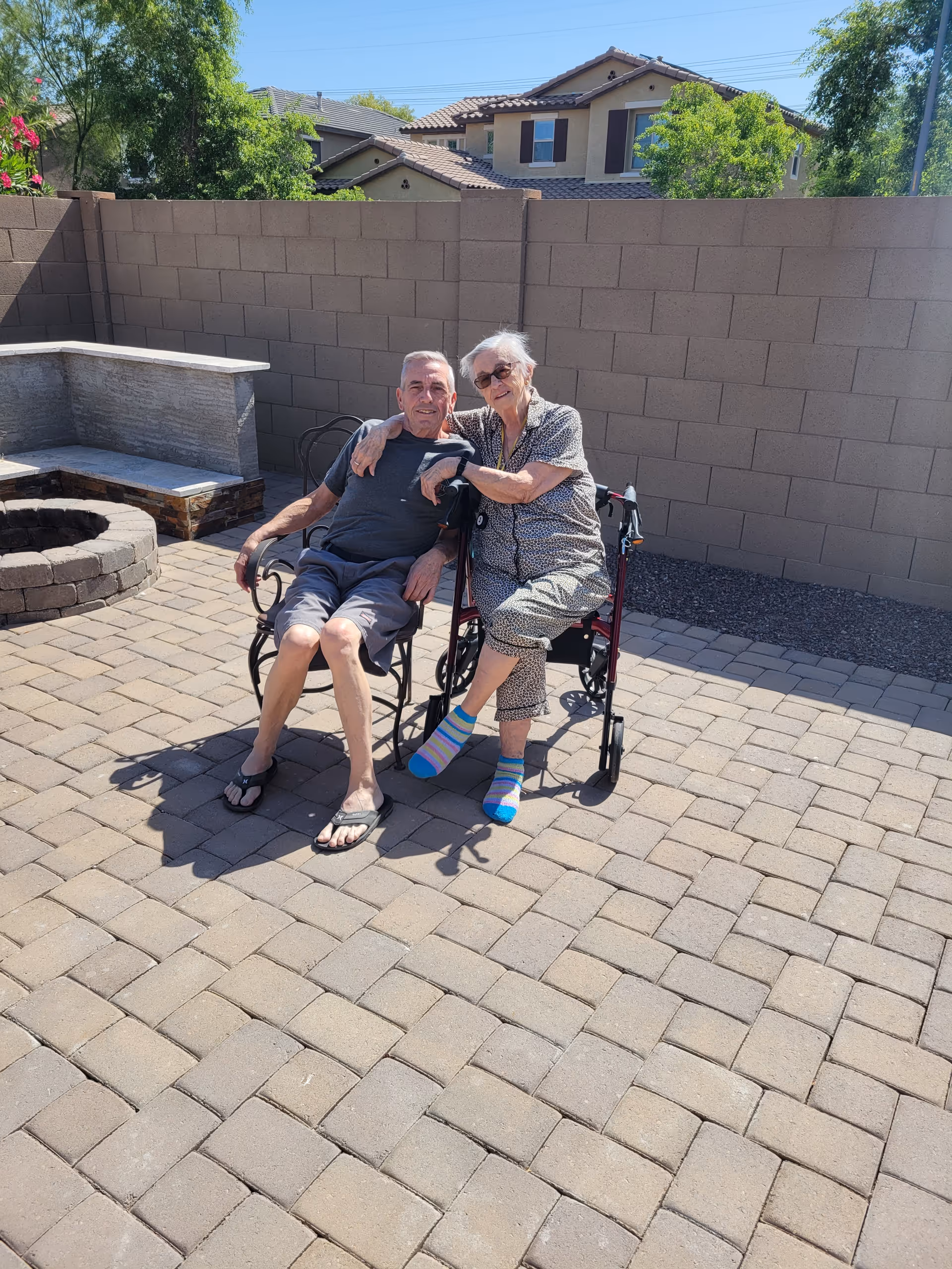 Two people seated together on a sunny paved backyard patio—one using a rollator/wheelchair—near a stone fire pit and a block wall with houses visible behind.