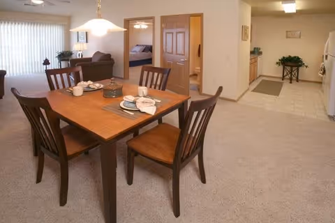 Interior view of a senior living facility showing a dining area with a wooden table set for four, a living room with a sofa and lamp in the background, a bedroom visible through an open door, and a kitchen area with appliances and cabinets.