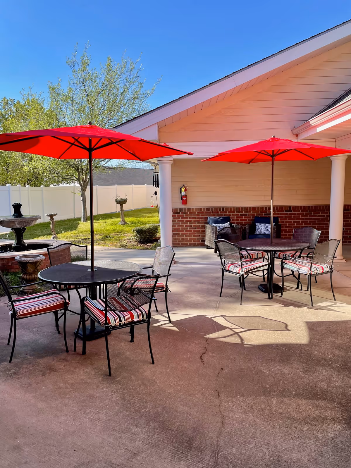 Outdoor patio area with two round tables, each shaded by a red umbrella. Each table is surrounded by four metal chairs with striped cushions. In the background, there is a brick and siding building with a covered porch area containing additional seating. The patio overlooks a grassy area with trees, birdbaths, and a white fence under a clear blue sky.