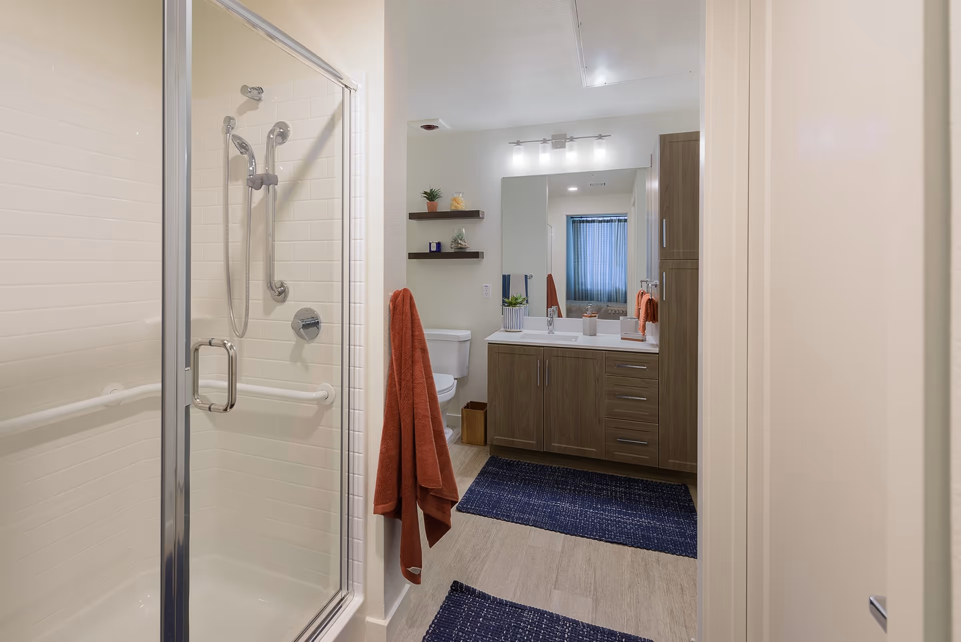 A modern bathroom featuring a glass-enclosed shower with grab bars, a wooden vanity with a large mirror and sink, a toilet, two dark blue rugs on the floor, and orange towels hanging on the wall and vanity. The room is well-lit with ceiling lights and a window with a blue curtain in the background.