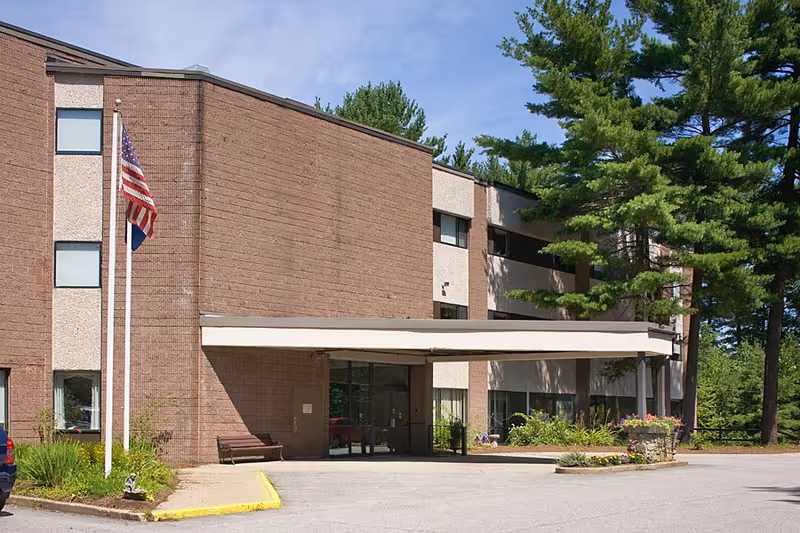 Front entrance of a brick senior living facility with a covered drop-off, two flagpoles, and surrounding trees.