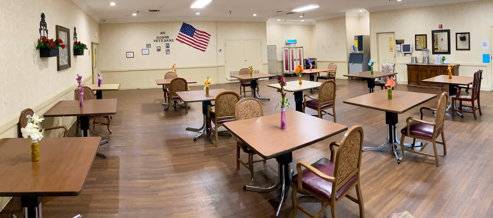 A spacious dining room with multiple wooden tables and chairs arranged neatly. Each table has a small vase with flowers. The room has wooden flooring, cream-colored walls, and an American flag on the far wall with a display that says 'We Honor Veterans'. There are framed pictures and wall decorations, and the room is well-lit with ceiling lights.