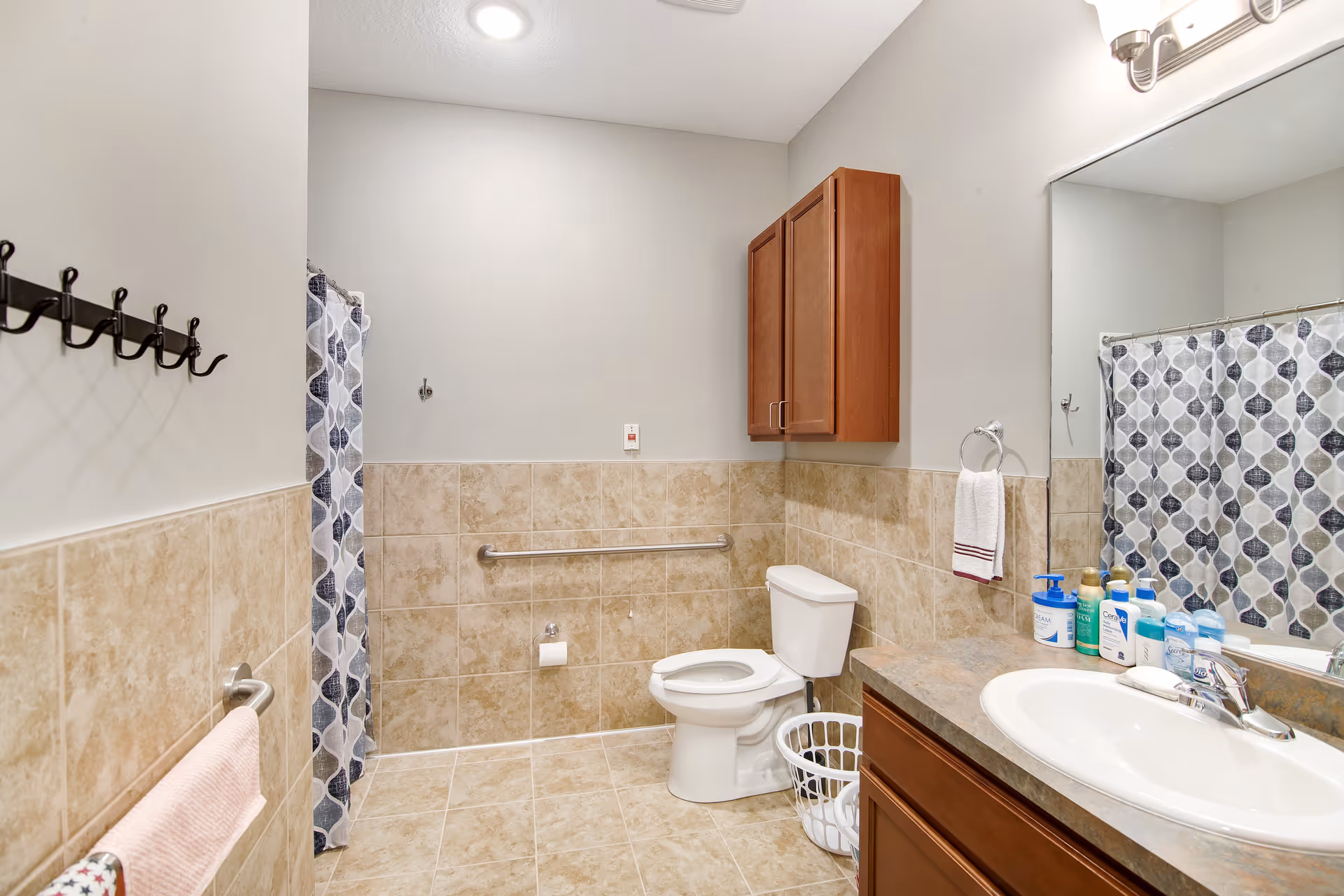 A clean bathroom with beige tiled walls and floor. There is a white toilet with a grab bar on the wall behind it, a white sink with a countertop and wooden cabinet below, and a large mirror above the sink. Various toiletries are arranged on the countertop. A shower with a patterned curtain is visible in the background. A towel rack with a pink towel is mounted on the left wall, and a wooden cabinet is mounted above the toilet.