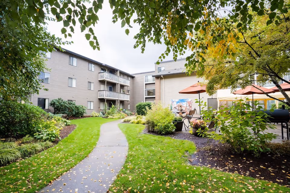 Outdoor view of Creekside Village Retirement Residence showing a paved walkway surrounded by green grass, bushes, and trees with some leaves turning yellow. The building has multiple floors with balconies, and there are patio tables with umbrellas on the right side.