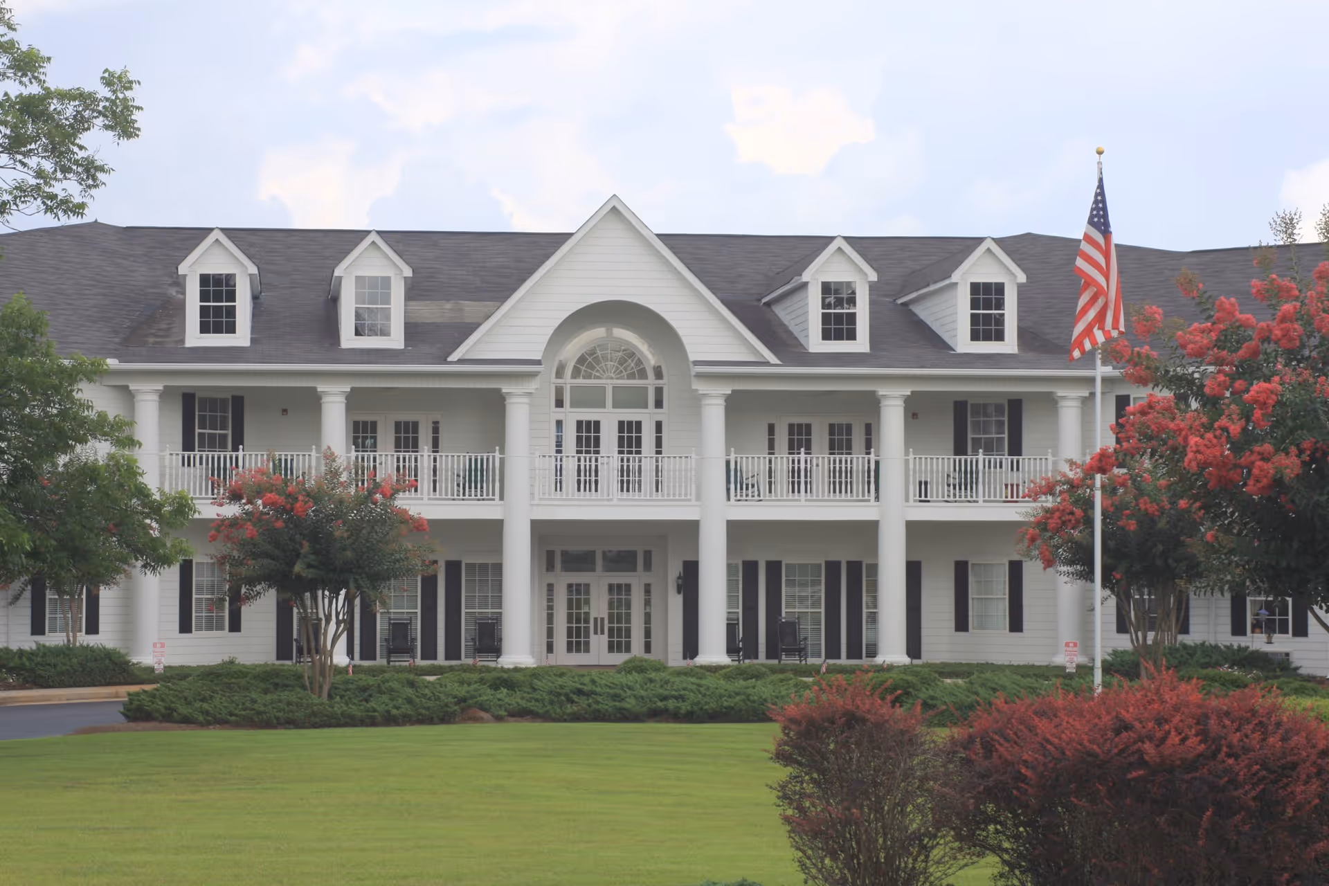 Front exterior of a large white two-story colonial-style senior living building with columns, balconies, an American flag, and landscaped lawn.