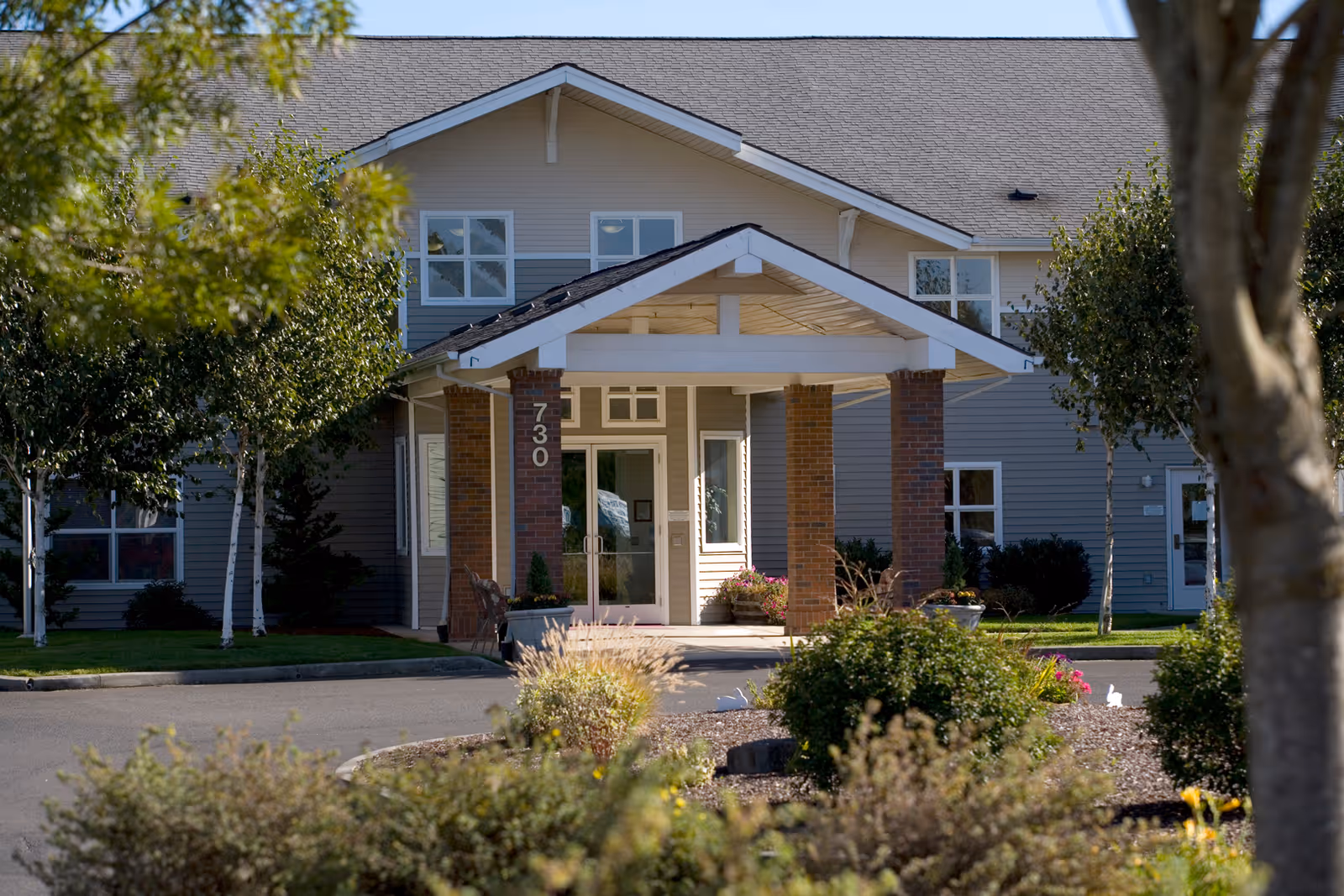 Front exterior view of a senior living facility named Avamere at Newberg, featuring a covered entrance with brick pillars, surrounded by trees and landscaping.
