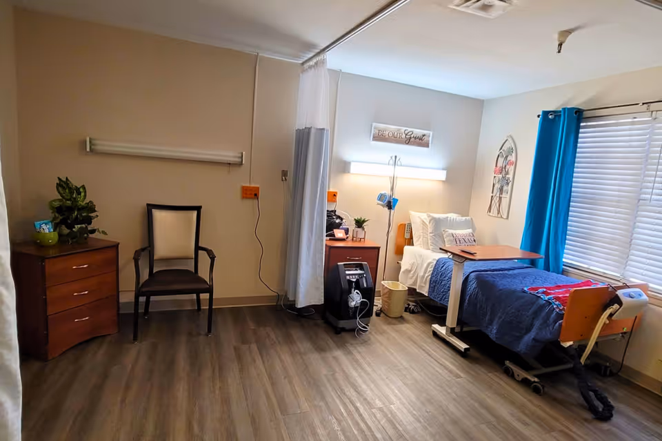 A clean and well-lit nursing facility bedroom with a hospital bed covered in blue bedding, a wooden bedside table with a lamp and small plant, a chair, a chest of drawers with a plant on top, a window with blue curtains, and a medical oxygen concentrator on the floor.
