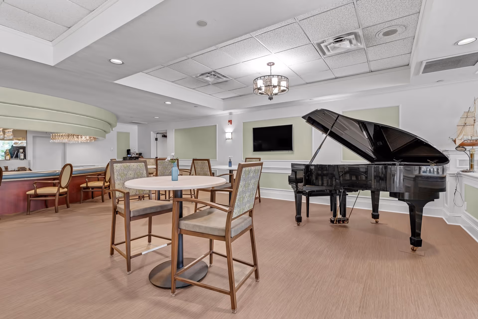 A spacious common area with a black grand piano, round tables with wooden chairs featuring patterned upholstery, a wall-mounted flat screen TV, and a bar area with additional seating. The room has light green and white walls, recessed lighting, and a chandelier.