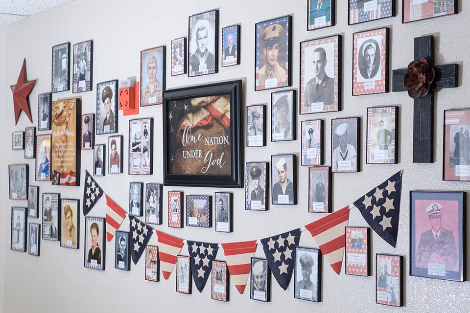 A wall decorated with numerous framed black and white and color photographs of military veterans, patriotic-themed bunting with stars and stripes, a red star, a wooden cross with a flower, and a framed sign that reads 'One Nation, Under God'.