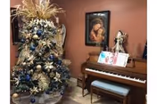 A decorated Christmas tree with blue, gold, and white ornaments and ribbons stands next to a wooden piano with a cushioned bench. On top of the piano are sheet music and a decorative angel figurine. A framed picture hangs on the wall above the piano.