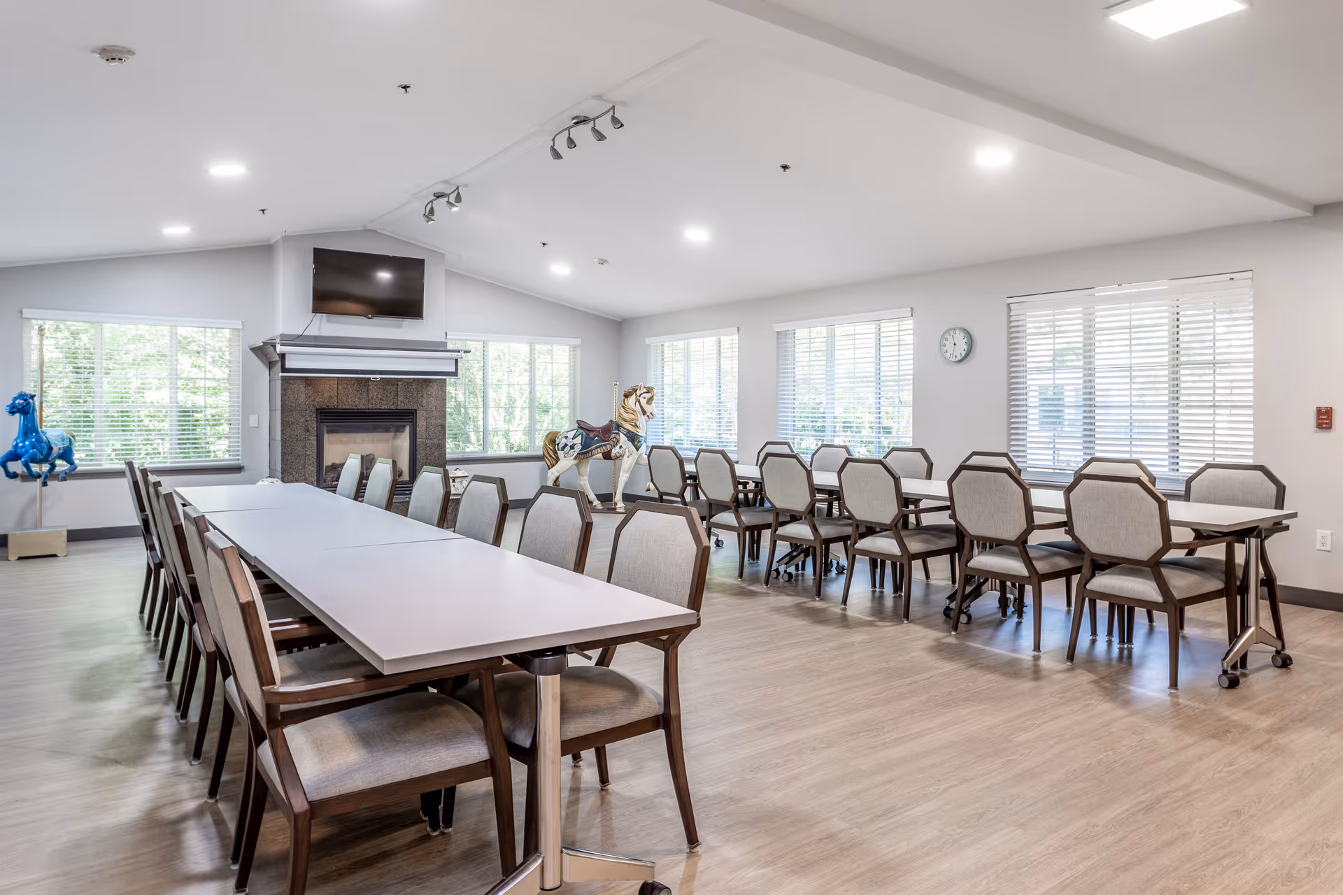 A spacious dining room with two long tables surrounded by multiple chairs. The room has large windows with blinds, a fireplace with a mounted TV above it, and two decorative carousel horse statues near the windows. The floor is wooden, and the ceiling has recessed lighting.