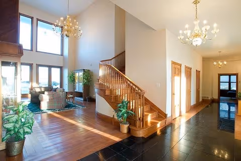 Bright and spacious interior of a senior living facility featuring a wooden staircase with a curved handrail, a sitting area with plaid upholstered chairs and a large window letting in natural light, potted plants, and chandeliers hanging from the ceiling.