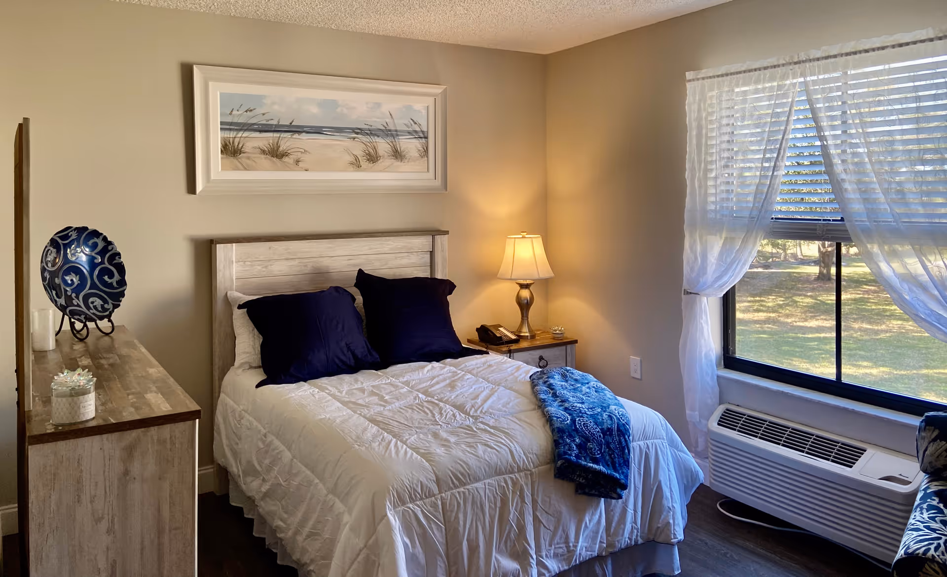 A cozy bedroom with a neatly made bed featuring white bedding and two dark blue pillows. A blue patterned throw blanket is draped over the foot of the bed. To the right of the bed is a wooden nightstand with a lamp and a phone. A large window with sheer white curtains lets in natural light, showing a view of green grass and trees outside. On the left side of the room is a wooden dresser with decorative items including a blue and white patterned vase and a small potted plant. Above the bed hangs a framed picture of a beach scene with sand dunes and grasses.