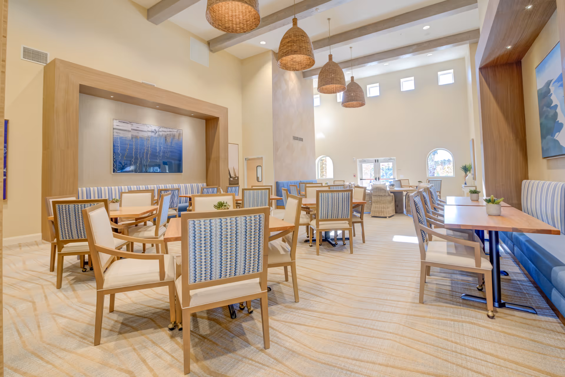 Bright, airy communal dining room with multiple wooden tables and upholstered chairs, pendant lights, and large windows.