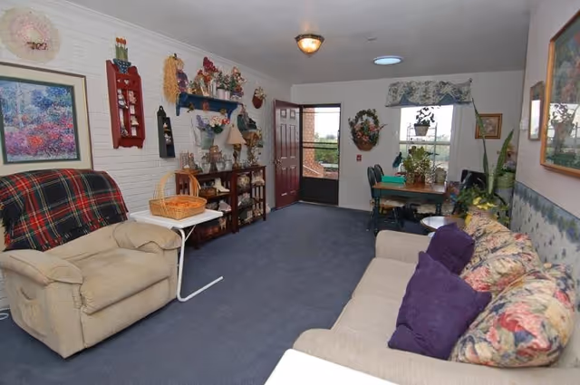 Cozy assisted-living common room with a sofa and armchair, decorative shelves, and a dining table by a window.