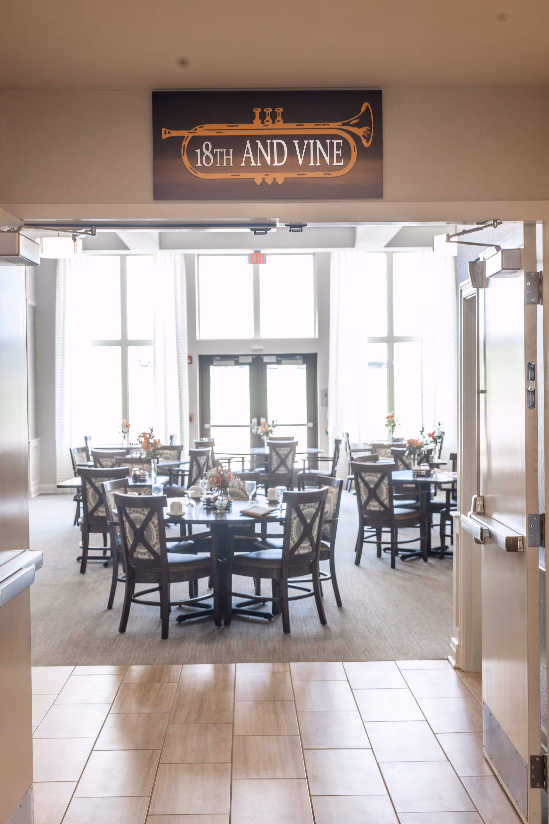 View through an open doorway into a bright dining room with several round tables and chairs arranged neatly. Each table is set with glasses, napkins, and small floral centerpieces. Large windows with white curtains allow natural light to fill the room. Above the doorway is a sign featuring a trumpet graphic and the text '18TH AND VINE'.