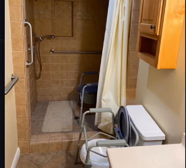 Accessible bathroom with a tiled walk-in shower, grab bars and shower chair, a toilet with a raised seat, and a wooden wall cabinet.