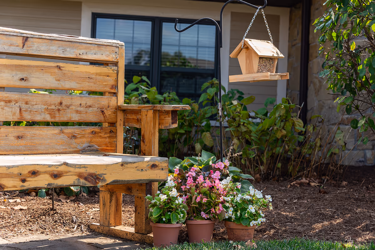 A wooden outdoor bench beside potted pink and white flowers and a hanging birdhouse in front of a building window.
