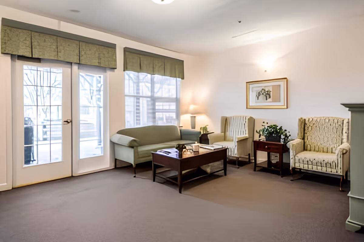 A cozy living room area with a green sofa, two patterned armchairs, a wooden coffee table, a side table with a plant, a lamp, and a framed picture on the wall. Large windows and glass doors with green valances allow natural light to fill the room.