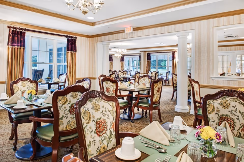 A bright and elegant dining room with multiple tables set for a meal. The tables have floral upholstered chairs, green placemats, folded beige napkins, cups, glasses, and silverware. The room features large windows with curtains, chandeliers, and decorative columns.