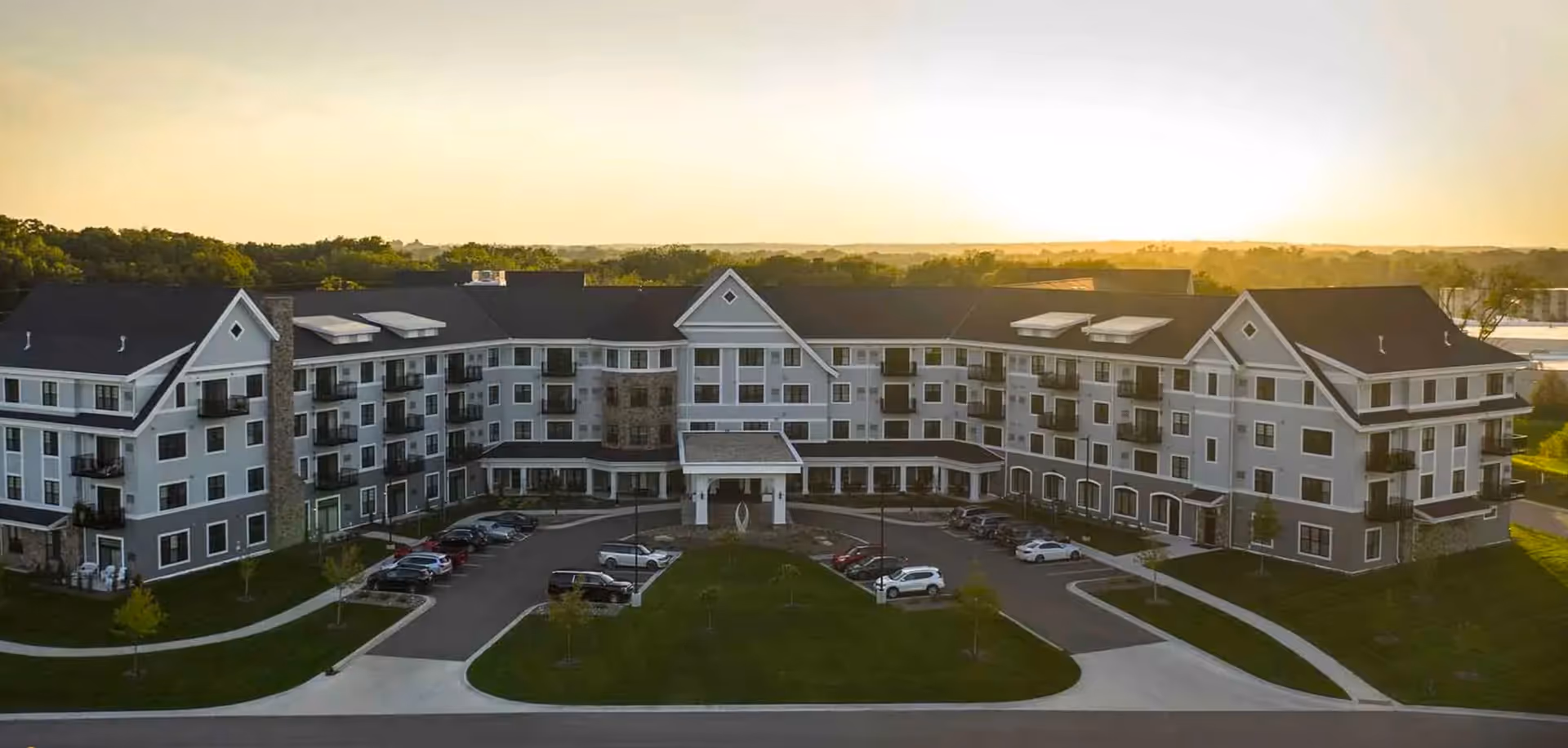 A large, multi-story senior living facility building with a U-shaped design, multiple balconies, and a covered entrance. The building is surrounded by a parking lot with several cars and landscaped green areas. The sun is setting in the background, casting a warm glow over the scene.