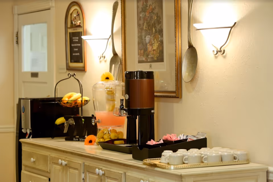 A beverage station in a senior living facility with a coffee dispenser, a container of pink lemonade with lemon slices and grapes, a basket of bananas, a tray with white coffee cups, and packets of sugar and creamer. The wall behind has decorative large spoon and fork hangings, a framed floral picture, and a menu board.