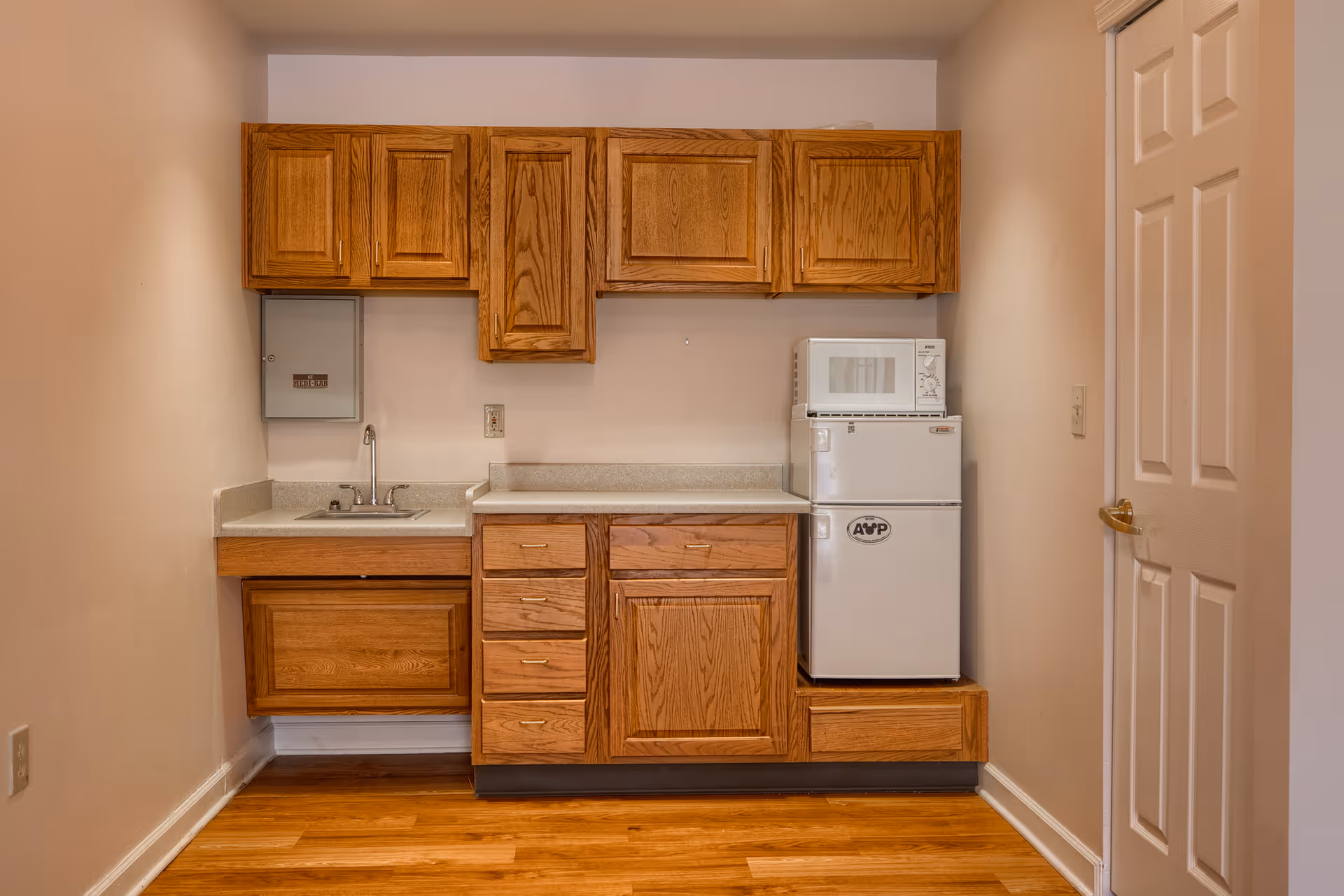 Small kitchenette with wooden cabinets, a countertop, a sink, a white mini refrigerator with a microwave on top, and a closed white door on the right side.