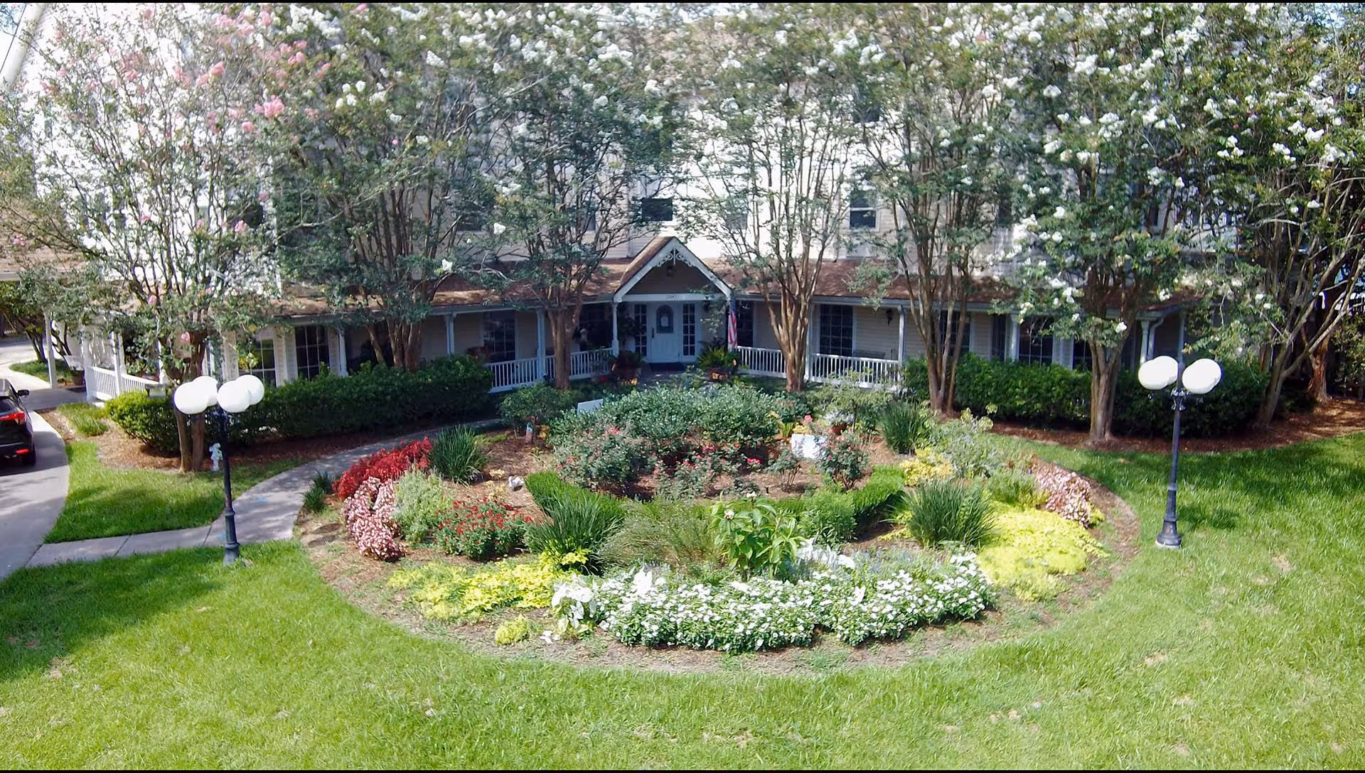 Front exterior view of St Francis Villa Assisted Living facility with a circular flower garden in the foreground, surrounded by green grass and trees with white and pink blossoms. The building has a porch with white railings and a peaked entrance.