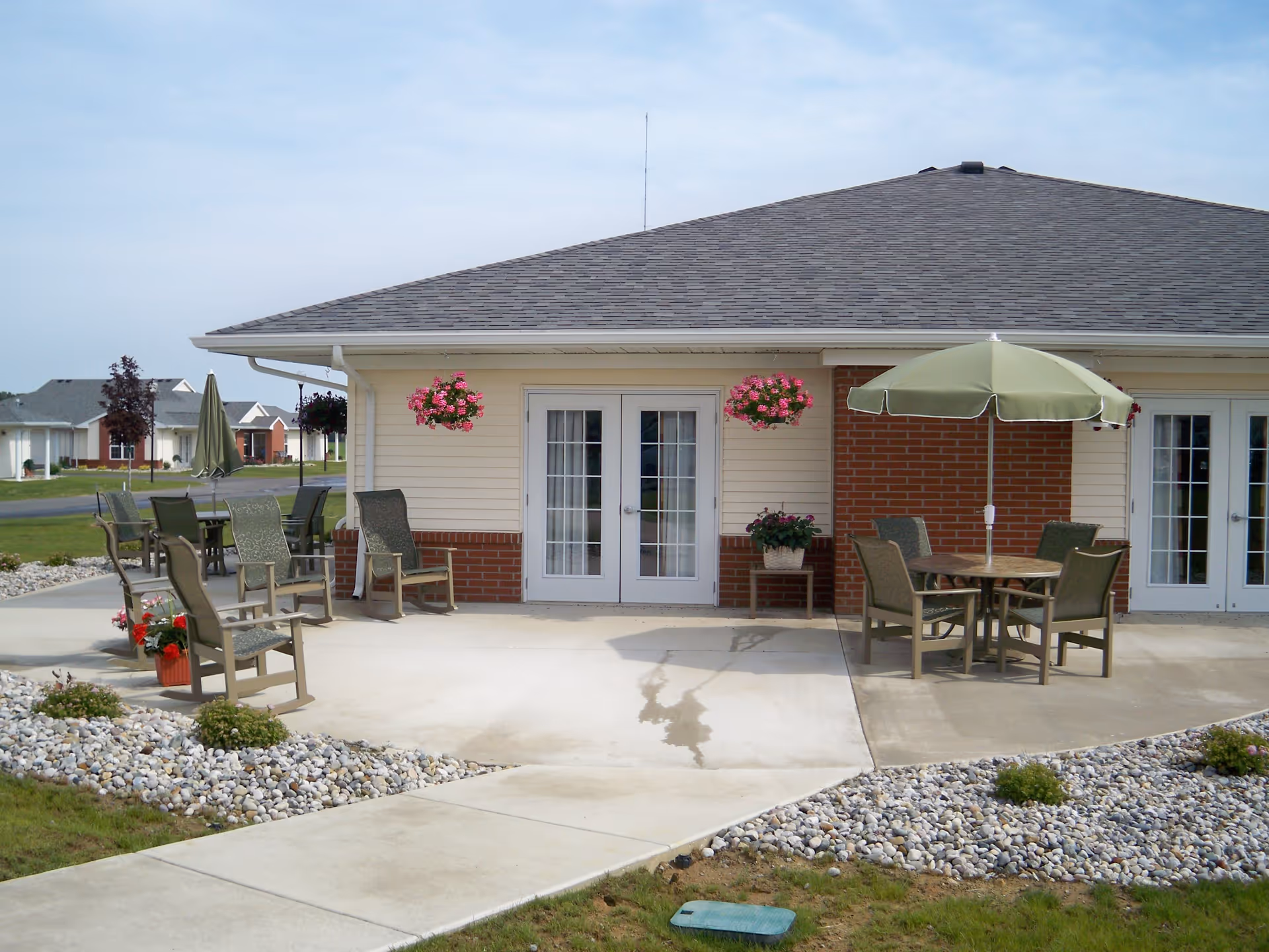 Patio outside a senior living building with seating, tables under umbrellas, hanging flower baskets, and double glass doors.