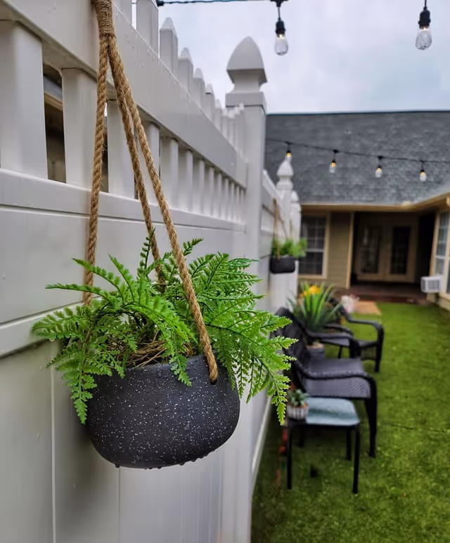 A hanging potted fern on a white fence overlooking a courtyard with chairs, string lights, and a building in the background.