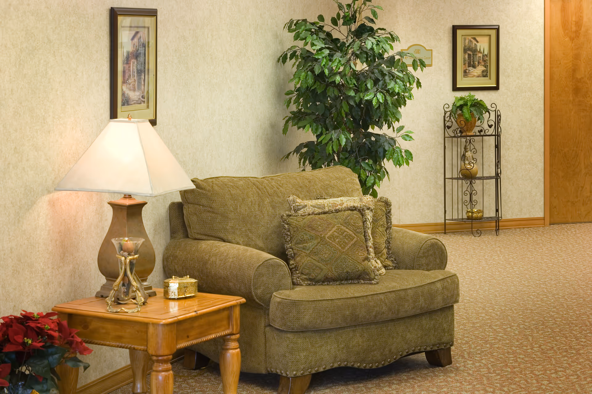 A cozy seating area in a senior living facility with a green upholstered loveseat adorned with two patterned cushions. Next to the loveseat is a wooden side table with a decorative lamp and small ornaments. The background features a tall green plant, a metal shelf with potted plants, framed artwork on the walls, and a wooden door.