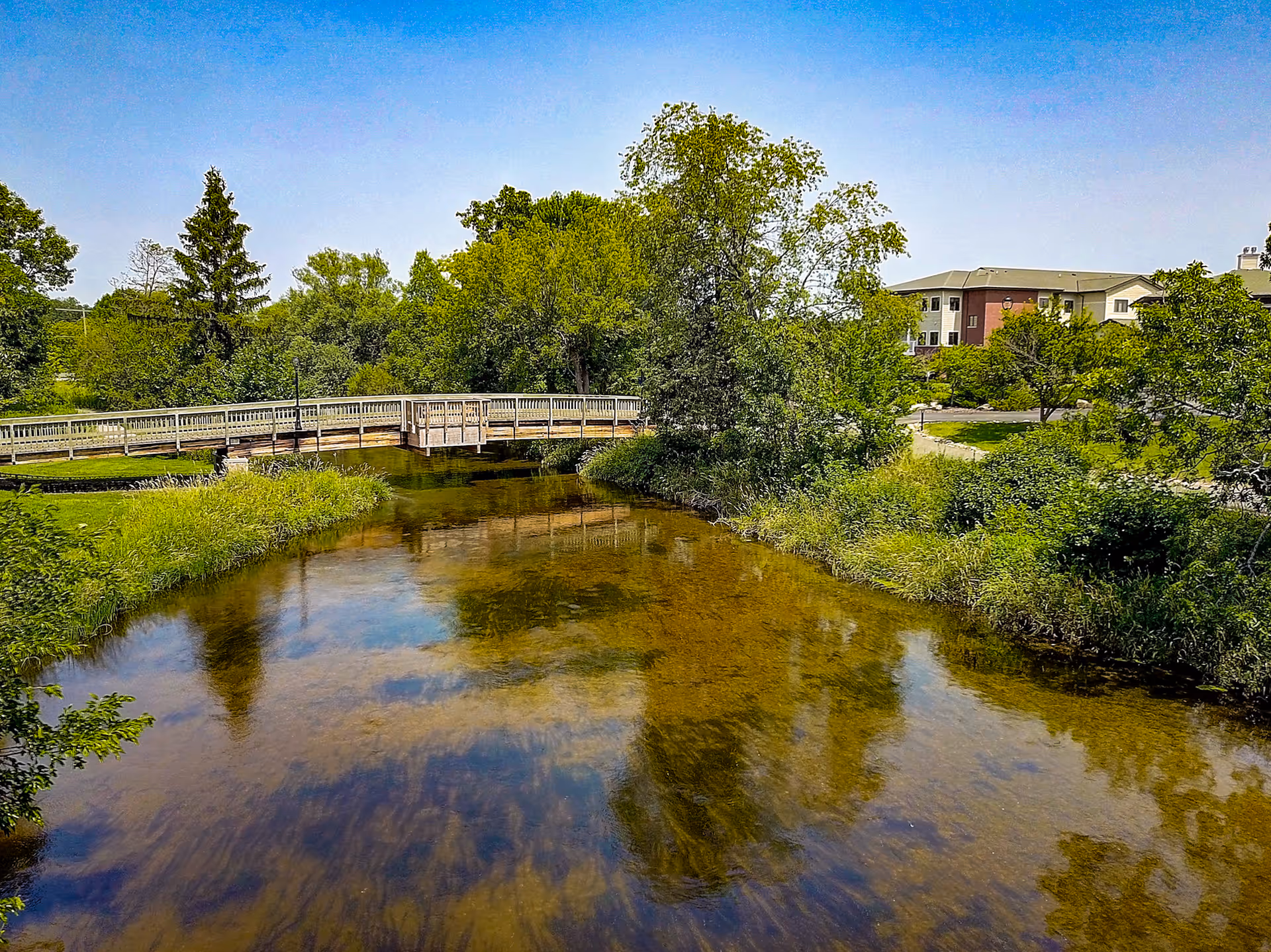 A clear, shallow river with reflections of trees and a blue sky. A wooden pedestrian bridge crosses the river, surrounded by lush green trees and grass. In the background, a multi-story building is partially visible behind the trees.
