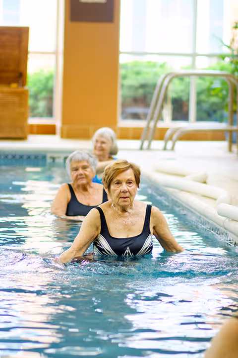 Three elderly women in swimsuits participating in water exercise in an indoor swimming pool with large windows and greenery visible outside.