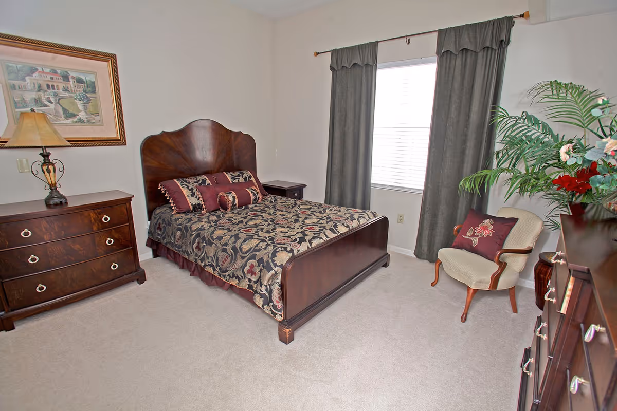A bedroom with a wooden bed frame and floral patterned bedding. There is a wooden dresser with a lamp and framed artwork on the wall to the left. A window with dark curtains is on the back wall, and a cushioned armchair with a decorative pillow is placed near a large green plant. The room has light-colored carpet and neutral walls.