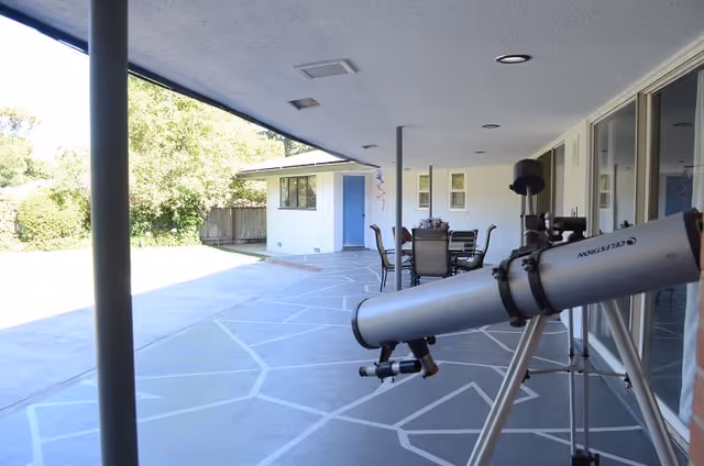 A covered outdoor patio area with a telescope in the foreground, a table with chairs in the background, and a small building with a blue door surrounded by greenery.