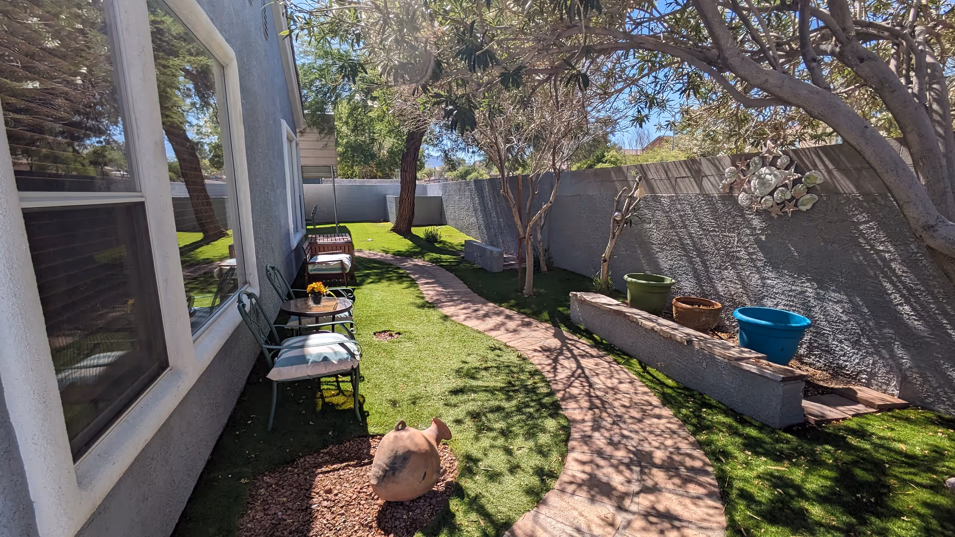 A narrow outdoor garden area with a curved stone pathway surrounded by green grass and trees. On the left side, there are two chairs with cushions and a small table with a flower pot. On the right side, there are several empty colorful plant pots on a raised concrete planter against a gray wall. The area is shaded by tree branches overhead.