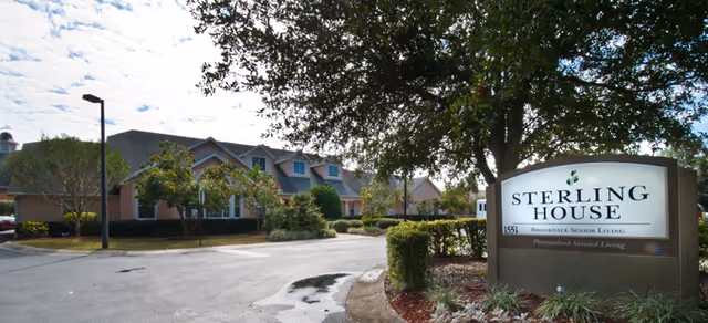 Front exterior of a single-story senior living building with trees, landscaping, a driveway, and a sign reading "Sterling House".