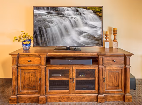 A wooden entertainment console with a flat-screen TV on top displaying an image of a cascading waterfall. The console has two drawers, two cabinets with wooden doors on the sides, and two glass-paneled cabinets in the center. On the left side of the console is a blue vase with yellow flowers, and on the right side are two decorative candle holders. The background wall is beige.