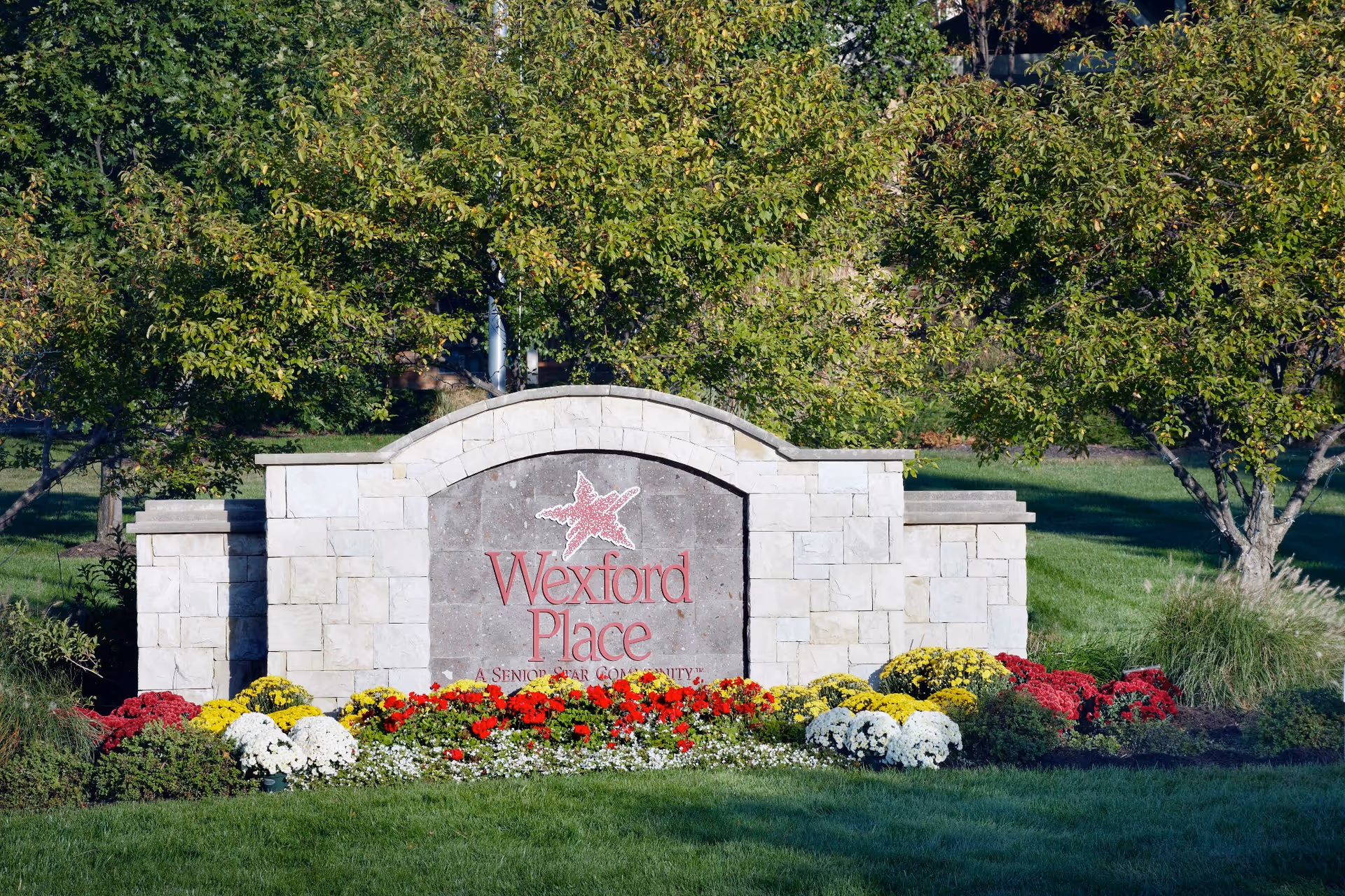 Stone sign for Wexford Place, a Senior Star community, surrounded by colorful flowers and green trees in a landscaped outdoor area.