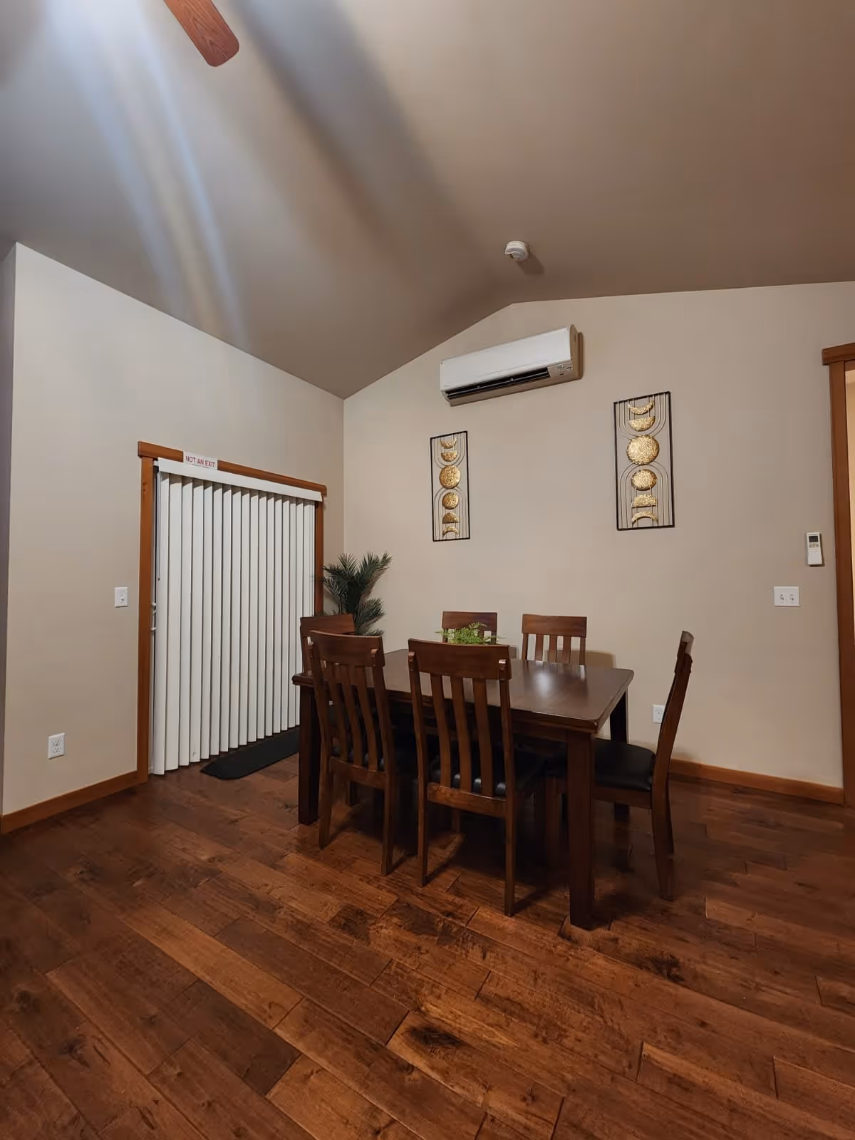 Wooden dining table with six chairs in a room with hardwood floors, vertical blinds, wall art, and a wall-mounted air conditioner.