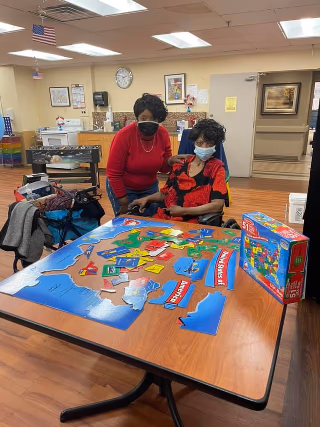 Two women wearing face masks are in a room with wooden flooring and beige walls. One woman is seated in a wheelchair at a table with a large colorful puzzle of the United States, while the other woman stands beside her with a hand on her shoulder. The room has a clock on the wall, framed pictures, and an American flag hanging from the ceiling.