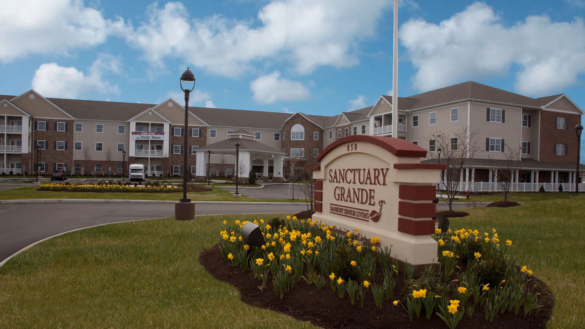 Exterior view of Sanctuary Grande, a senior living facility, showing a large three-story building with a covered entrance, surrounded by green lawns and flower beds with yellow flowers under a partly cloudy sky.