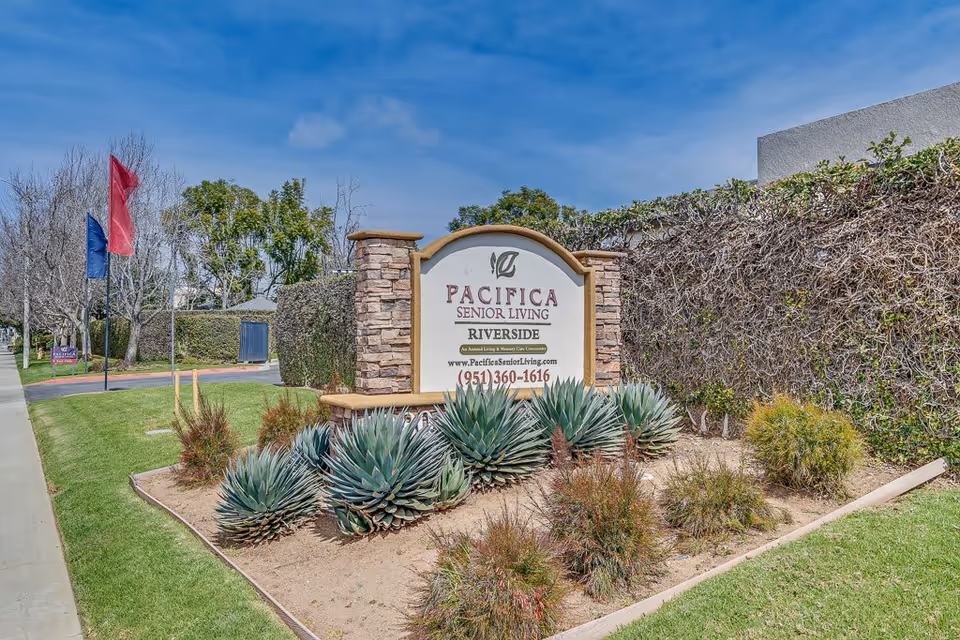 Outdoor view of the entrance sign for Pacifica Senior Living Riverside, surrounded by landscaped plants and greenery with a clear blue sky in the background.