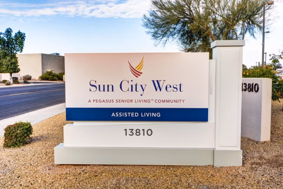 Large outdoor entrance sign for Sun City West Assisted Living displaying the address 13810 on gravel landscaping beside a road.